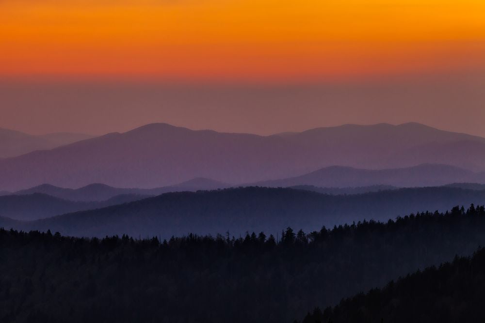 A horizontal photograph of the Great Smoky Mountains at sunset. The sky is a vibrant gradient, starting with a bright, fiery orange at the very top, blending downwards into softer oranges and then into a dusky rose-purple before meeting the mountain range. The mountains themselves are depicted in layers, creating a beautiful sense of depth and atmospheric perspective. The closest ridge, in the foreground, is the darkest, a deep silhouette of a forest with individual tree shapes visible. Beyond this, successive ridges fade into lighter shades of deep blue and purple, becoming progressively hazier and less defined as they recede into the distance.