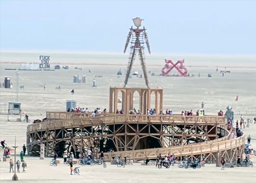 A screenshot of the live stream from Burning Man 2025 showing the base of the man and two art pieces, including one that says "F* You Elon"