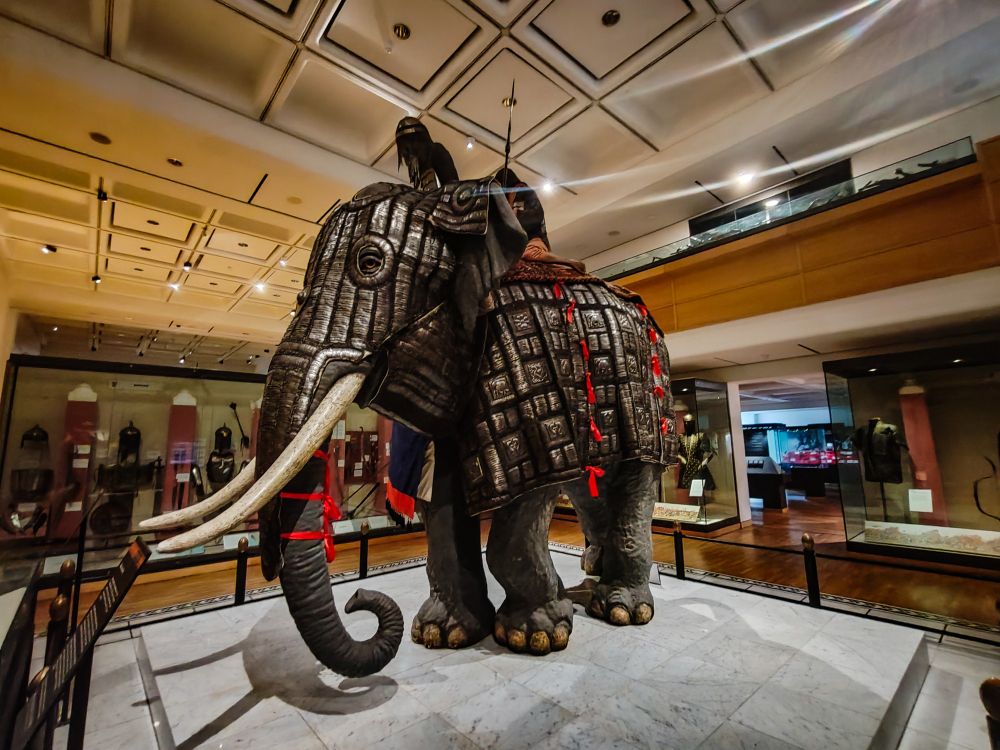 This photo showcases a remarkable exhibit at the Royal Armouries Museum in Leeds: a fully armoured elephant. The elephant is standing on a marble platform, surrounded by display cases featuring various artifacts. The elephant's intricate armor is made of metal and adorned with intricate details. Its tusks are visible, and it is wearing red and white cloths around its neck. The high ceiling of the museum is adorned with architectural details and recessed lighting. The overall atmosphere is awe-inspiring and historical, highlighting the unique and fascinating nature of this exhibit.