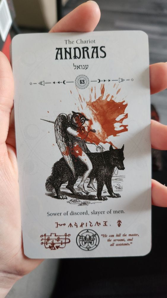 a tarot card, the chariot, featuring the demonic entity Andras from Travic McHenry's demon tarot deck. Based on the Ars Goetia / Lesser Key of Solomon. The image is of a humanoid body with large wings and the head of an owl, wielding a sword and riding on the back of a wolf. The card is white, but it features a bright red blood splatter across the surface. Binding sigils cover the card, with the warning words "he can kill the master, the servant and all assistants" marked in the bottom right corner.