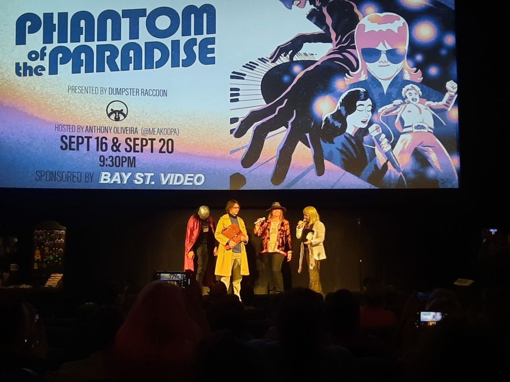Anthony Oliveira and audience members dressed as characters from Phantom of the Paradise against the backdrop of the show poster projected on the screen. 