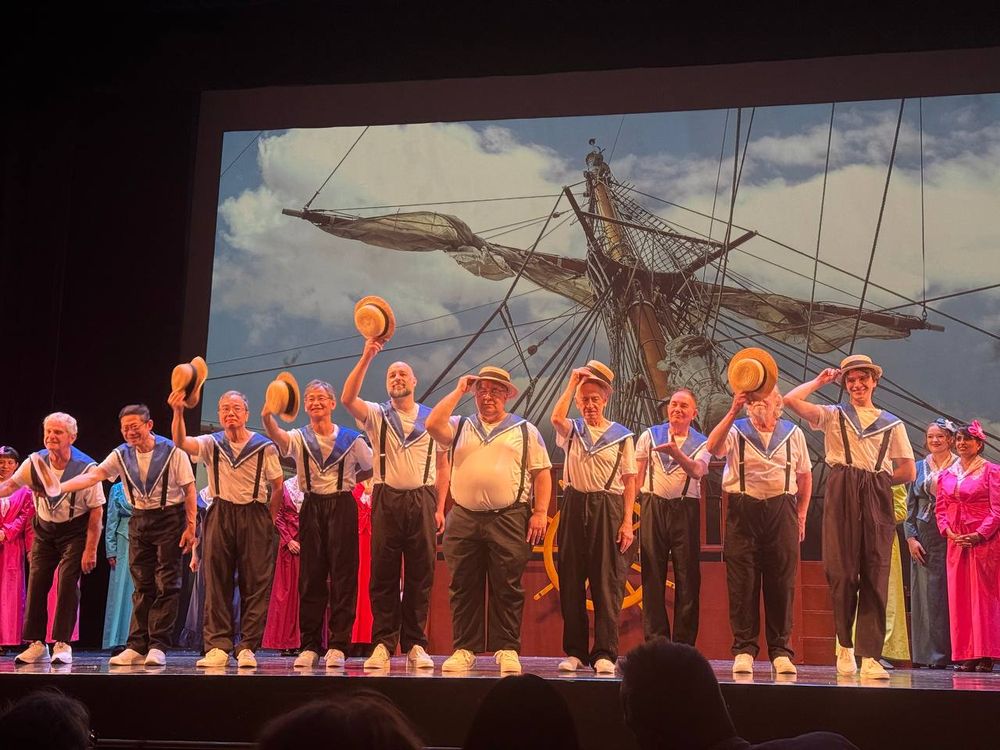 A line of men dressed as sailors in the men's chorus of HMS Pinafore lifting their hats to bow at the end of the show.