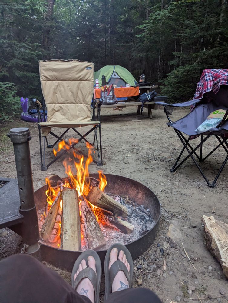 Wood burning in a fire pit with someone's feet next to it. Behind is a tent, picnic table, and empty chairs. 