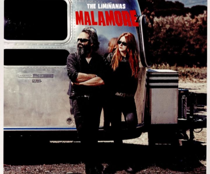 Cover des Albums Malamore von The Limiñanas. Ein schwarz gekleidetes Paar (er links stehend mit verschränkten Armen, schwarzem Vollbart und Sonnenbrille, sie rote lange Haare, ebenfalls Sonnenbrille; beider Blicke nach links gewandt) steht an einen silbernen Wohnwagen mit getönten Scheiben gelehnt auf Kies; im Hintergrund Hecken.