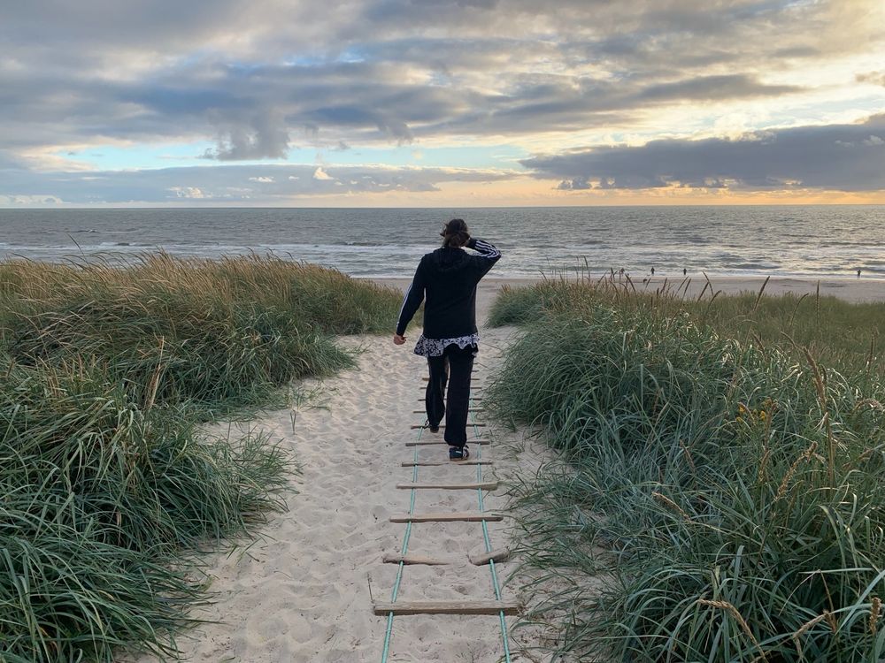 Eine Frau geht einen Sandweg eine Düne hinunter ans brausende Meer, links und rechts wächst Strandhafer; am Himmel i
zerklüftete Wolken lassen vereinzeltes Blau durchblitzen. Alles ist in leicht goldenes Licht getaucht.