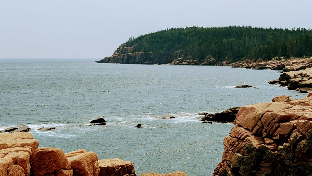 The picture of the ocean with rocks on the side. A rock formation covered in trees in the distance. Cloudy skies, muted, Waters, calm