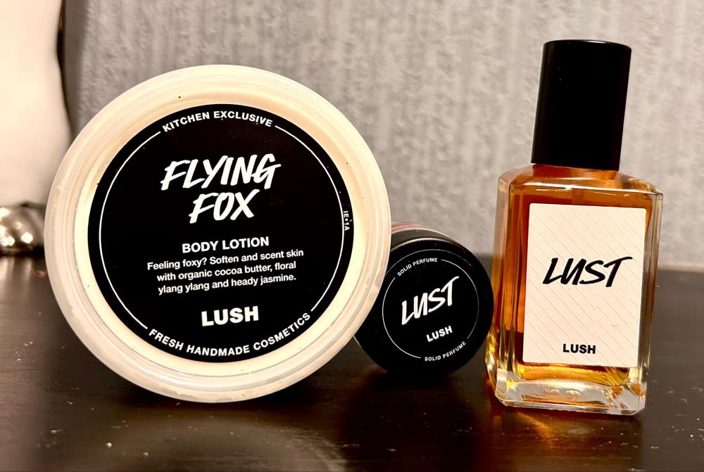 A short rectangular clear glass bottle full of some translucent amber yellow liquid with a metallic black lid and a white label that has Lush Lust Perfume written in bold black writing next to a short cylindrical glass jar with a black plastic lid and a white label that has Lush Lust Solid Perfume written in bold black writing and a short cylindrical black plastic tub with a black label that has Lush Flying Fox Body Lotion written in bright white writing on it on a black wooden cabinet. 