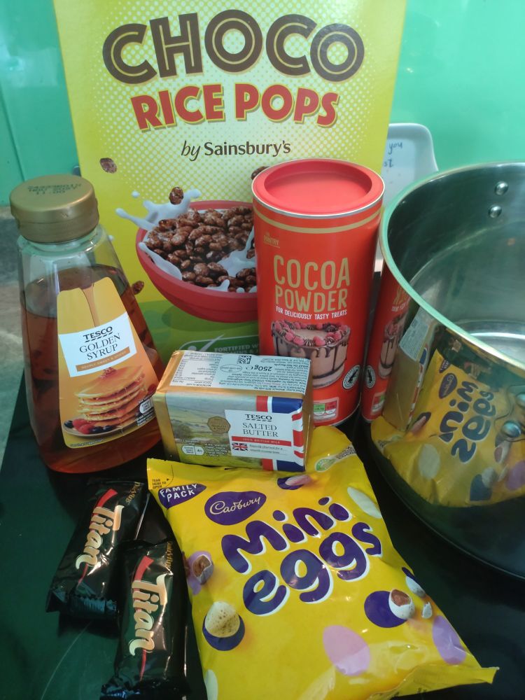 A silver pot, a box of chocolate rice pops cereal, tub of cocoa powder, bottle of golden syrup, block of salted butter, Aldi mars bars, and a large bag of mini eggs 