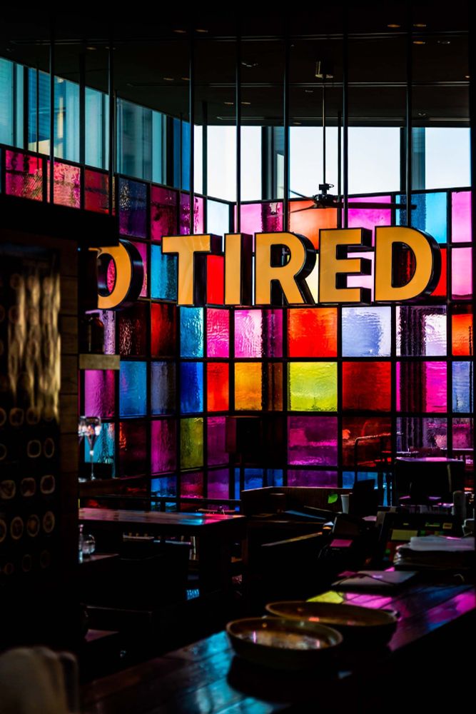 A darkened bar, cafe or restaurant. We can just about see a counter top with plates, tables and chairs but the room is dark. There is a panel on the wall with squares of coloured glass with the evening/morning light coming through: red, yellow, pink, blue and pale purple. 

Letters in front of the glass squares are in orange and spell: TIRED