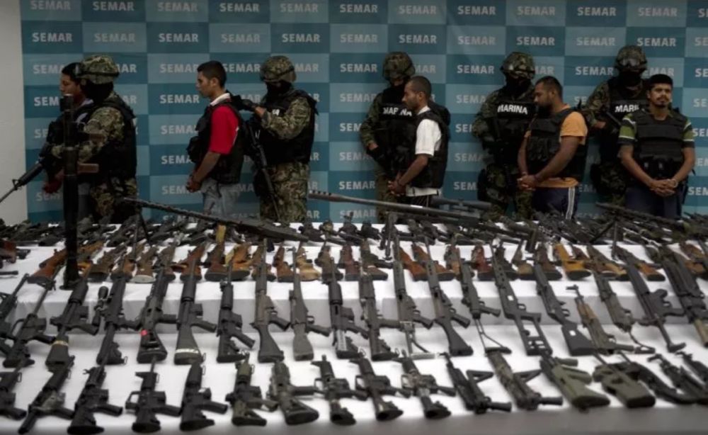 Mexican Naval Officers displaying arrested cartel members stopped amidst an arms trafficking operation. In front of the line of officers and cartel members are various rows of firearms and rifles found in said operation.