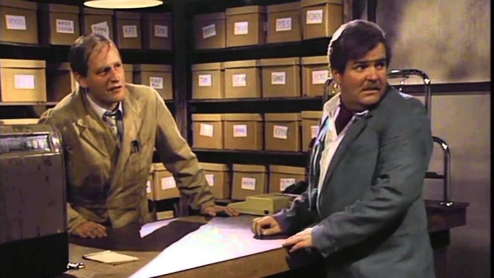 A sales assistent and a customer in a shop, that sells "stuff" from brown cardboard boxes.