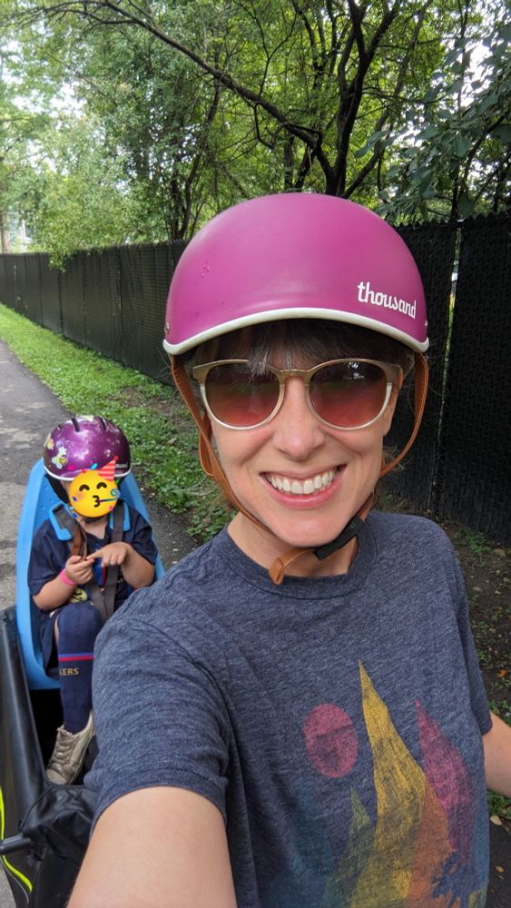 A selfie of Cassie on a bike wearing a purple helmet. A child also wearing a purple helmet is in a bike seat behind her.