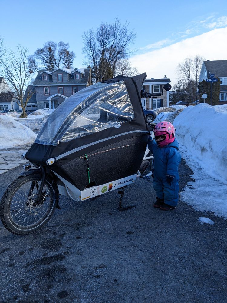 An Urban Arrow e-bakfiets on a driveway between snowmounds. A small child wearing a snowsuit, balaclava, helmet and sunglasses.