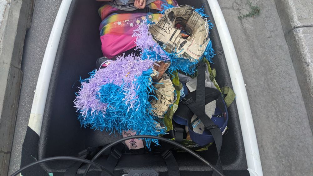 The bucket of a cargo bike as seen from above. A kid sits in the front across from two halves of a pinata.