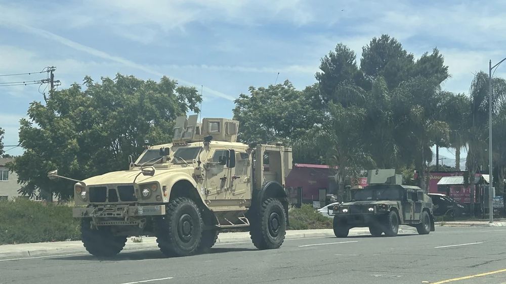 A military Humvee is dwarfed by an assault vehicle painted in desert tan, featuring armored gun turrets on the roof, armored exterior, and bristling with menace. It's tires are as tall as the humvee behind it. It is strange and disturbingly out of place in a scene that would normally be described as a pleasant neighborhood.