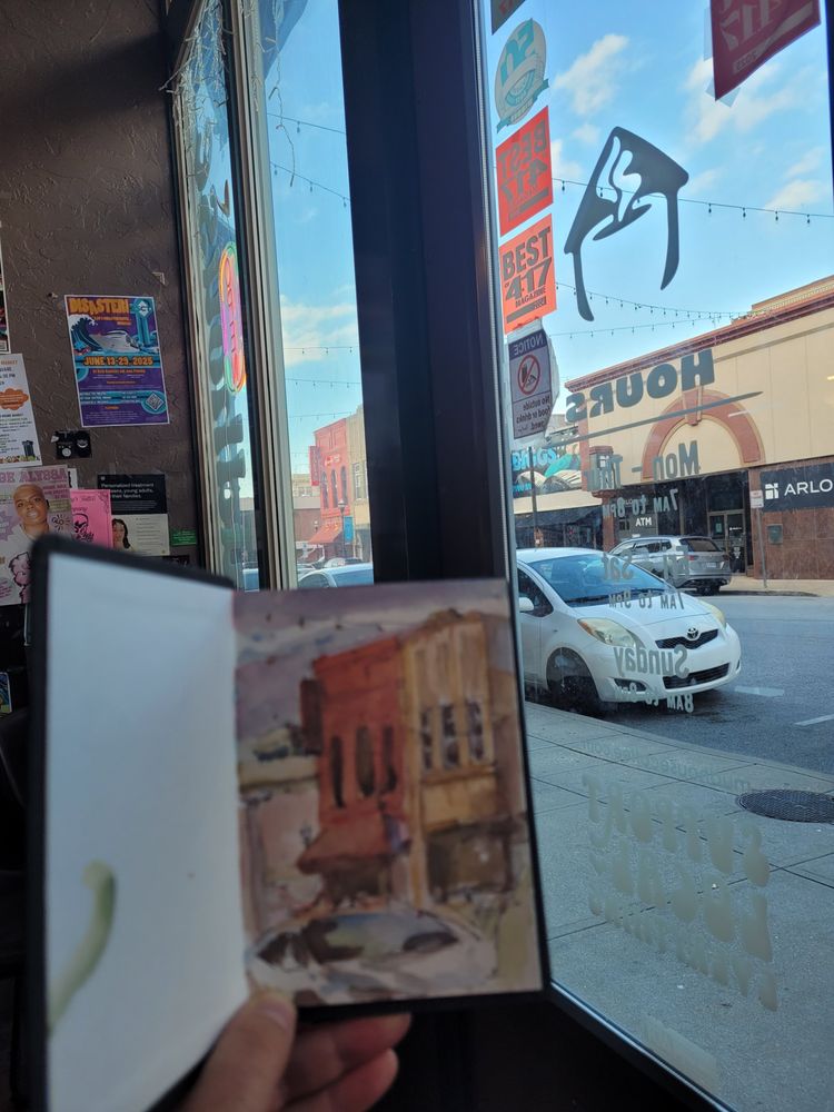 a watercolor and ink sketch being held up in front of the streetscape that it depicts -- a sun-kissed downtown building with some cars parked on the street