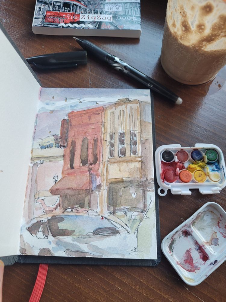 a sketchbook on a counter, open to a watercolor and ink sketch of a small, mid-west downtown streets cape. a small, toy watercolor palette meant for kids, the packaging for the new hahnemule sketchbook, a pen and travel paintbrush are next to the book