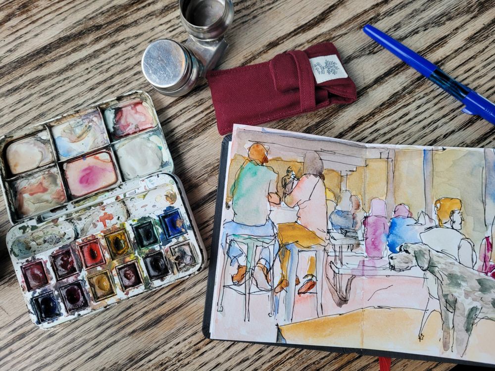 A top down view of an open sketchbook, watercolor palette and pair of water cups on a table, as well as a pack of travel paintbrushes in a carrying case.
The sketchbook has a colorful watercolor and ink depiction of a crowd of people on a restaurant terrace, with a fiddle player in the far background.