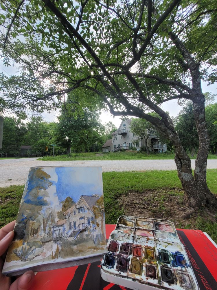 A large but run-down house in the background, surrounded by mature trees on a country road. The foreground shows a messy palette of watercolor paints and the small 2-toned sketch of the house and trees behind.