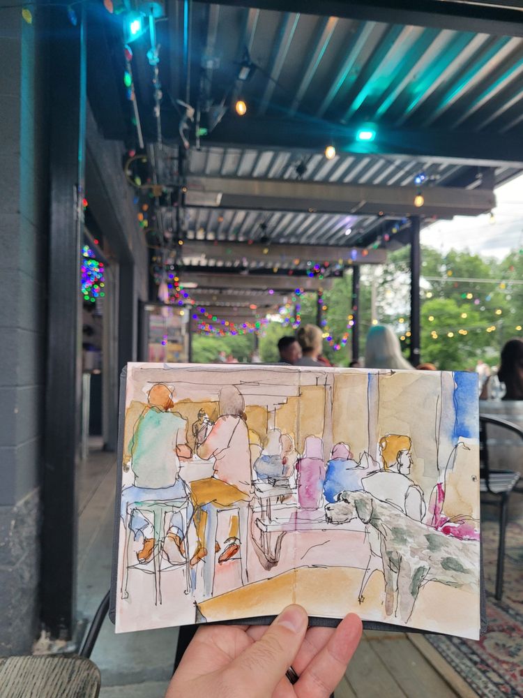 A top down view of an open sketchbook, watercolor palette
a sketchbook is held up and open in front of a restaurant terrace full of people.
The sketchbook has a colorful watercolor and ink depiction of a crowd of people on a restaurant terrace, with a fiddle player in the far background.