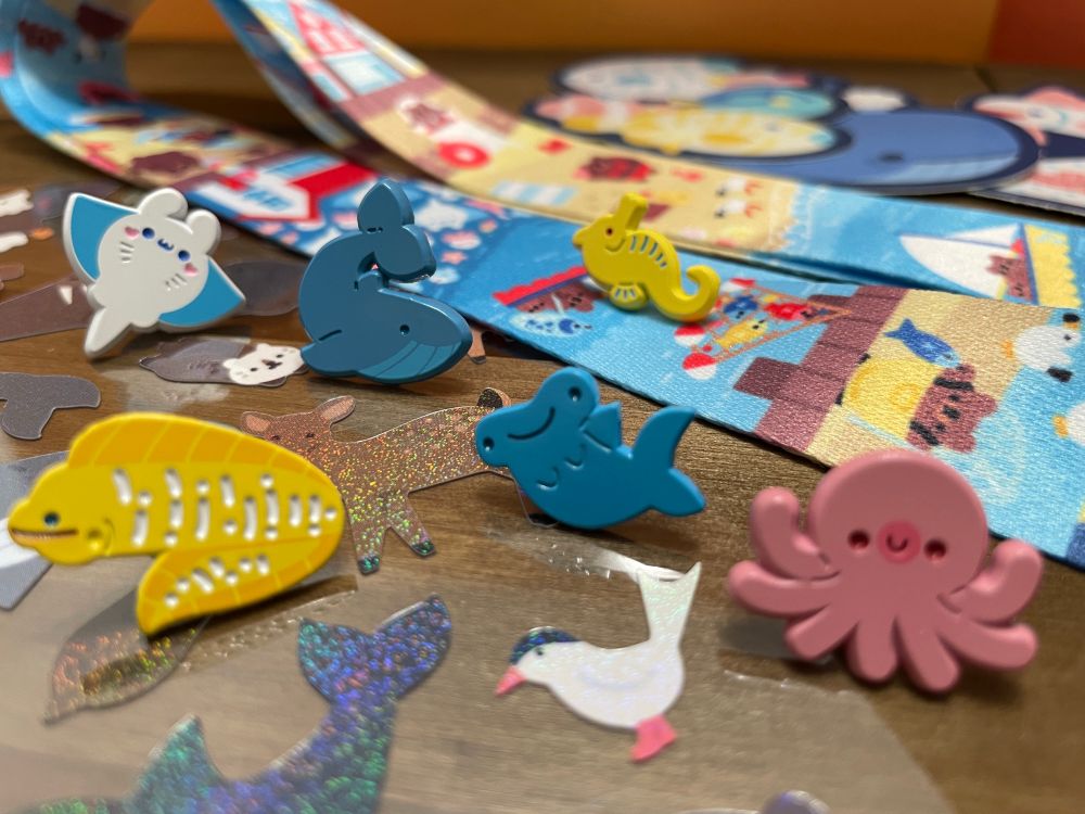 Photograph of a glittery sticker sheet, soft dyed enamel pins, large vinyl stickers, and a lanyard which all depict cute animals. Most of the animals have something to do with the ocean. 