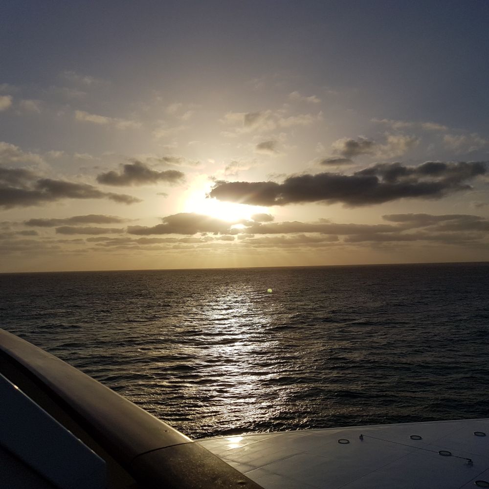 Blick vom Balkon eines Kreuzfahrtschiffes Richtung Sonnenauf- oder untergang. Das Meer ist relativ ruhig die Sonne scheint durch die Wolken. 