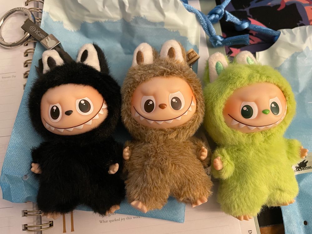 Three mini Labubus, one pitch black, one a mousey brown, and one chartreuse green. The green one has an impressive side-eye.