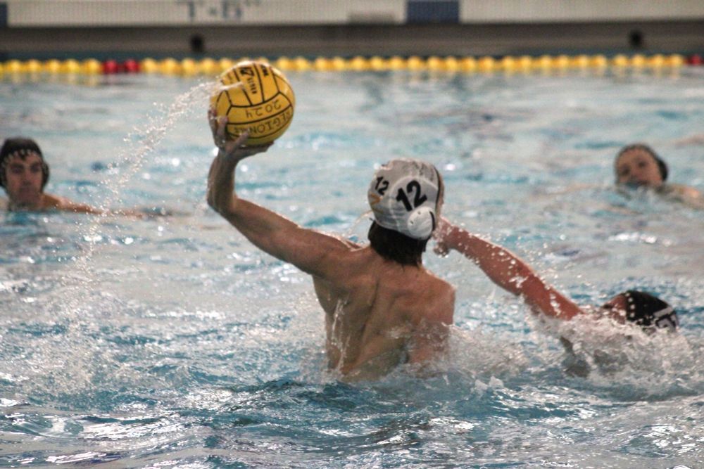 A male high school water polo player wearing a white cap #12 is rising up from the water holding a polo ball in preparation for a shot. The defender is swimming toward him with an arm ready to block. 