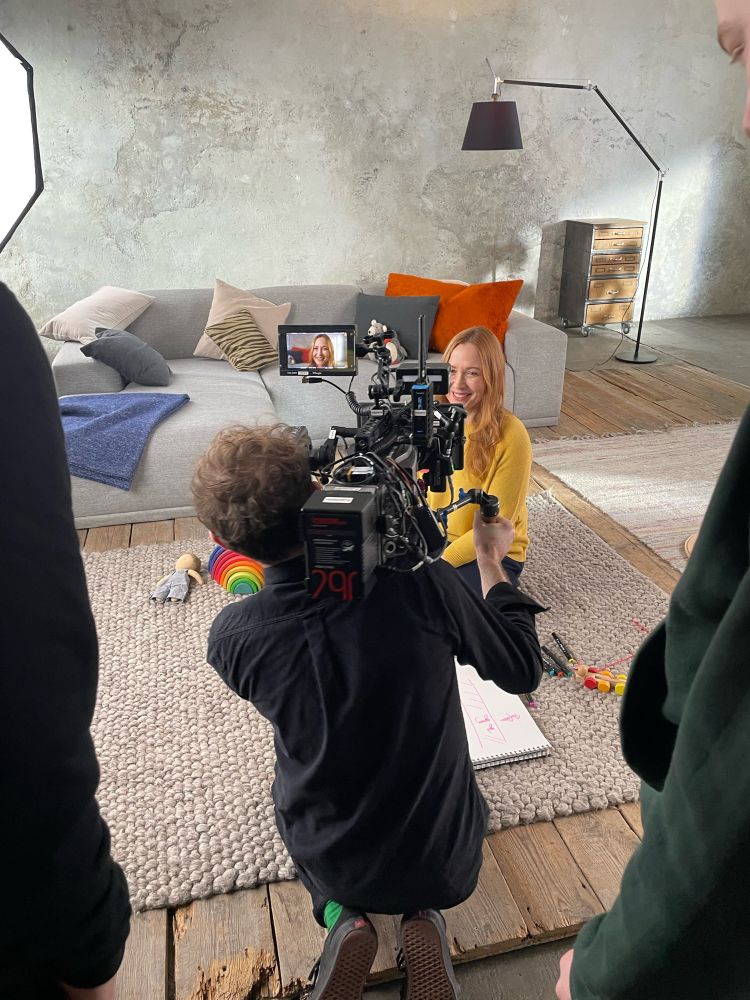 Foto von Susanne Mierau am Filmset mit Kameramann, Susanne sitzend auf dem Fußboden in gelbem Pullover vor einem Sofa mit Spielzeug