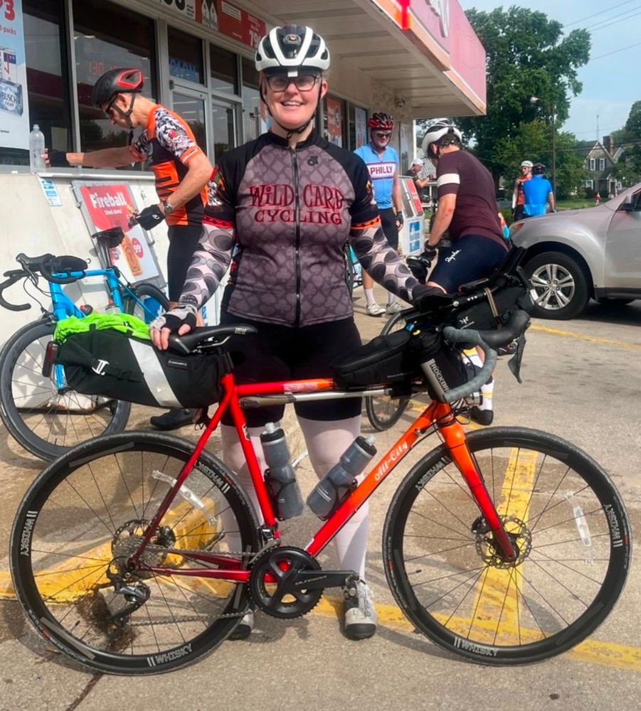 Smiling woman wearing cycling clothes holding a red and orange road bike in front of her. The bike is sporting a lot of bags in the back and front. 