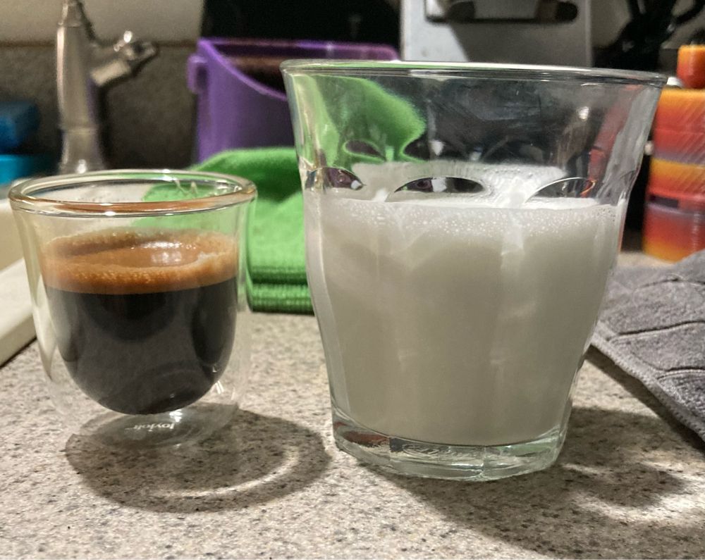 On the left, a small glass of dark brown espresso.9! The right, a taller glass 3/4 full of foamed milk. There are some snippets of two microfiber towels, a purple knockbox, and the espresso machine in the background.