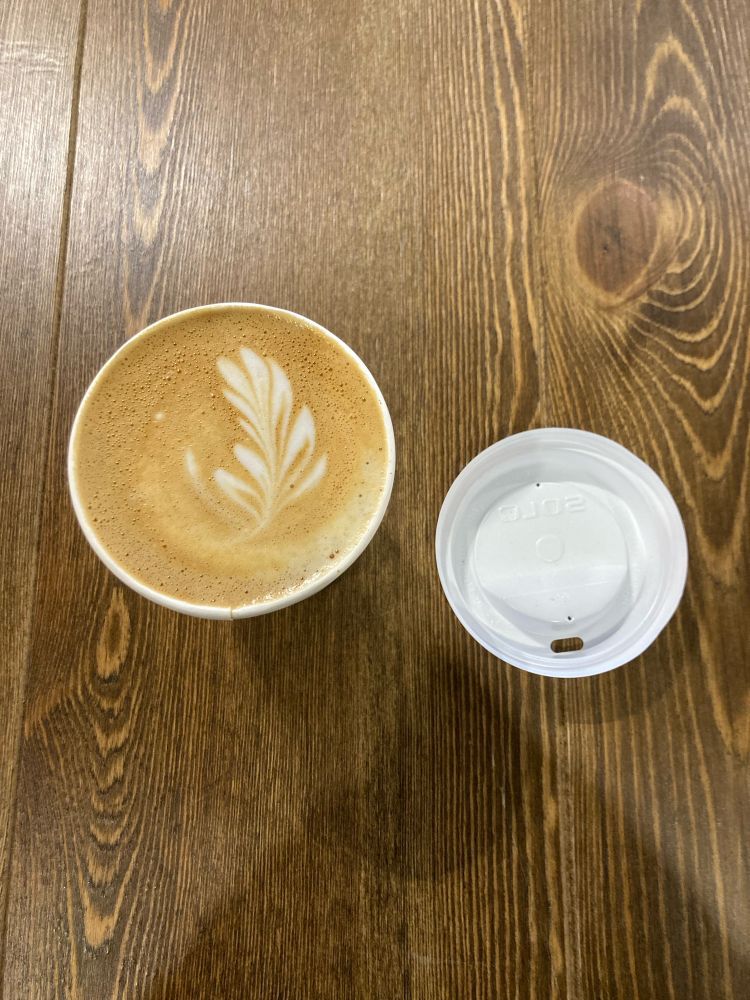 A picture of a cappuccino in a to-go up, with a lid to the right. The cappuccino foam is in the shape of a leaf.