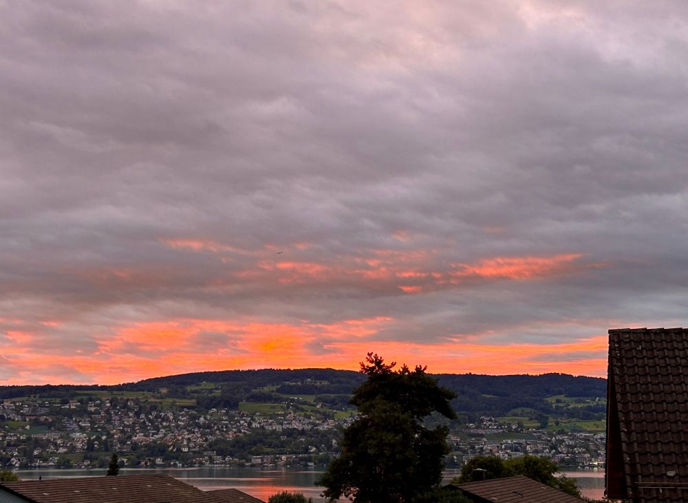 Morgendämmerung über einem See mit bewaldeten Hügeln und Häusern am Ufer. Der Himmel ist größtenteils wolkenverhangen, doch zwischen den grauen Wolken leuchten orange-rote Streifen des Sonnenaufgangs. Im Vordergrund sind Dächer und ein Baum zu sehen.