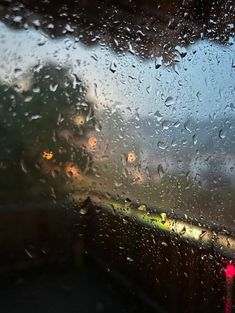 Ein Blick aus einem Fenster, das von vielen Regentropfen bedeckt ist. Im unscharfen Hintergrund erkennt man verschwommene Häuser, Bäume und Lichter in verschiedenen Farben. 
