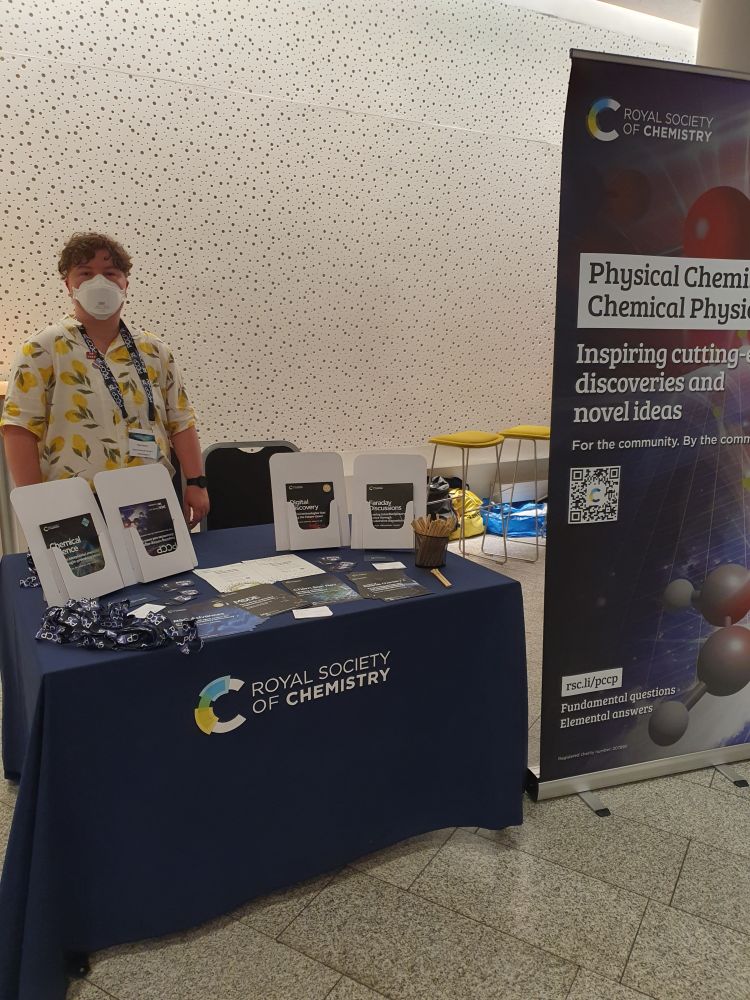 An R-S-C exhibition booth featuring a selection of leaflets promoting RSC journals as well as a large pop up banner celebrating the 25th anniversary of P-C-C-P. Behind the booth stands Bee, a white nonbinary person wearing a white shirt with lemons printed on it.