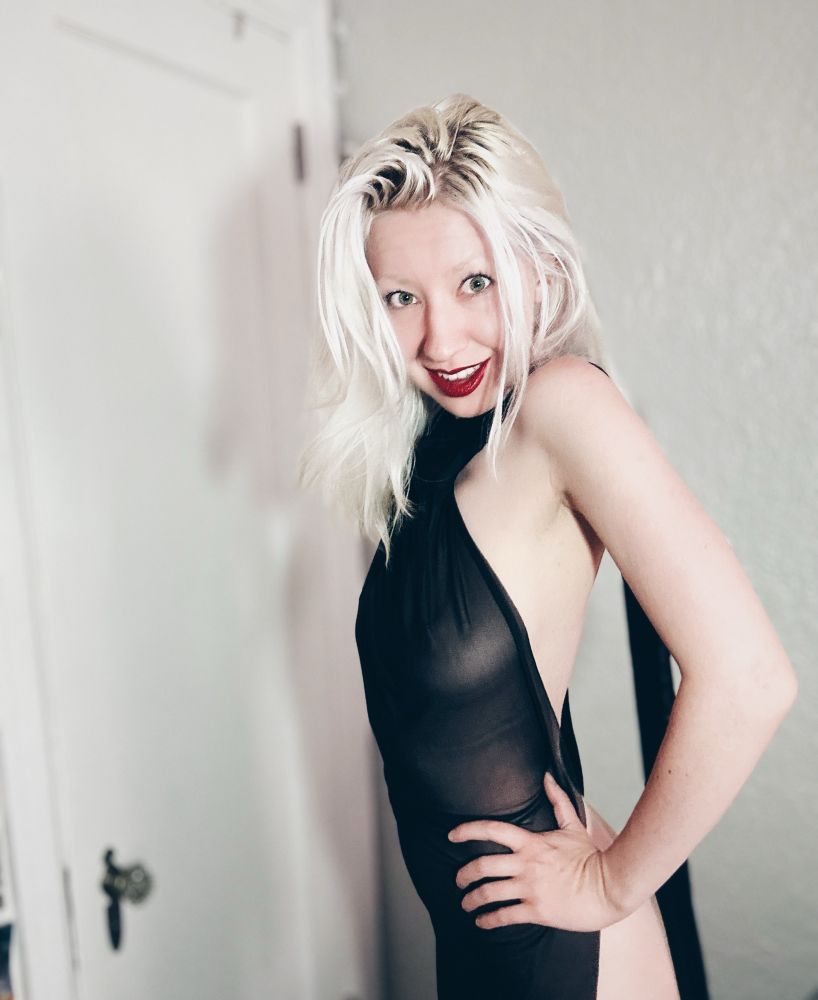 Valentine wearing a sheer black dress and red lipstick. He has her hands on her hips and there’s a door in the background 