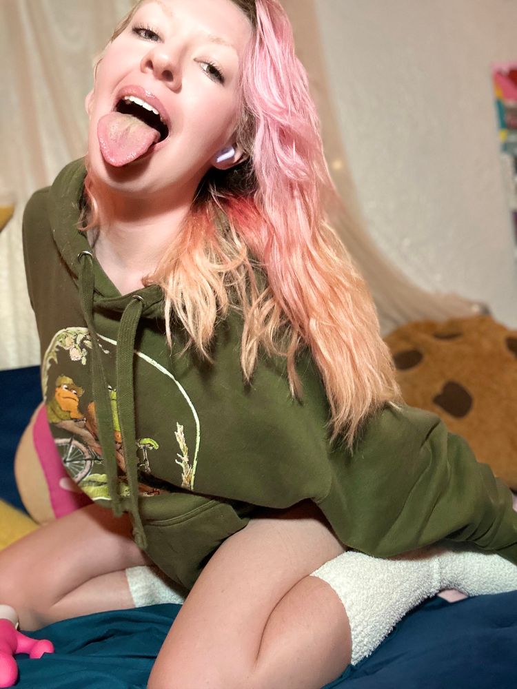 Valentine, kneeling on her couch next to a dildo, she is wearing white socks and a green frog and toad hoodie. She is sticking out her tongue.