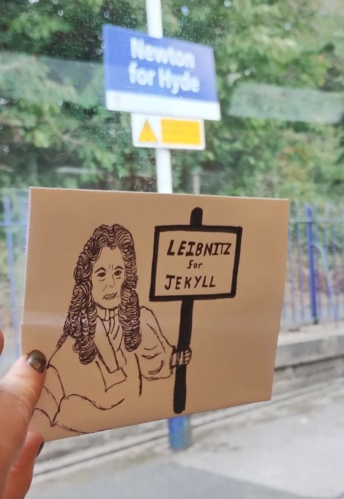 A photo taken through the window of a train. The name of the station "Newton for Hyde" can be seen on a sign. Inside the train a piece of paper is held up to the window with a sketch of a bewigged gentleman holding up a sign saying "Leibnitz for Jekyll"