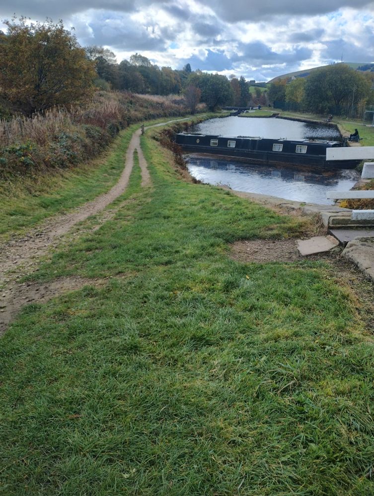 Looking down from the top of a canal lock to a canal basin that has been blocked by a black canal boat diagonally. The surrounding area is green with autumnal trees. The sky is a mix of blue with grey and white clouds. 