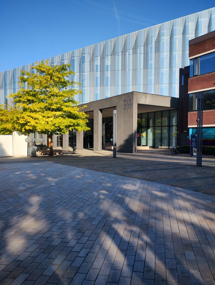 A modern glass sided building with a concrete awning, in front of which is a green leaved tree.