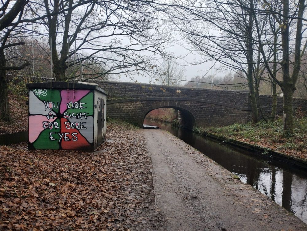 A canal running under a stone bridge. The towpath is on the left of the canal. Left of the towpath is a square utility building with bright graffiti saying "you are a sight for sore eyes" on it. The day is grey.
