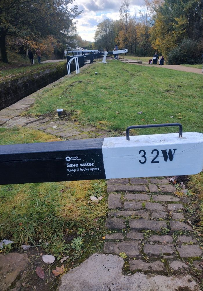 A canal lock bar with 32W on it and a note to save water-keep two locks apart. The background shows green space on an autumn day.