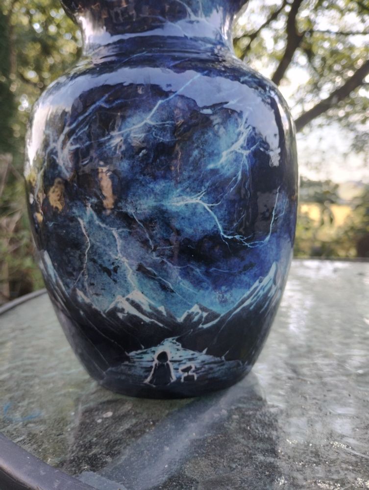 A vase featuring  a gorgeous dark blue nebula design resembling a lightning storm over a road leading through mountains. A hooded figure and a small robot are looking down the road. The vase is sitting outside on a glass table with green fields and trees in the background.