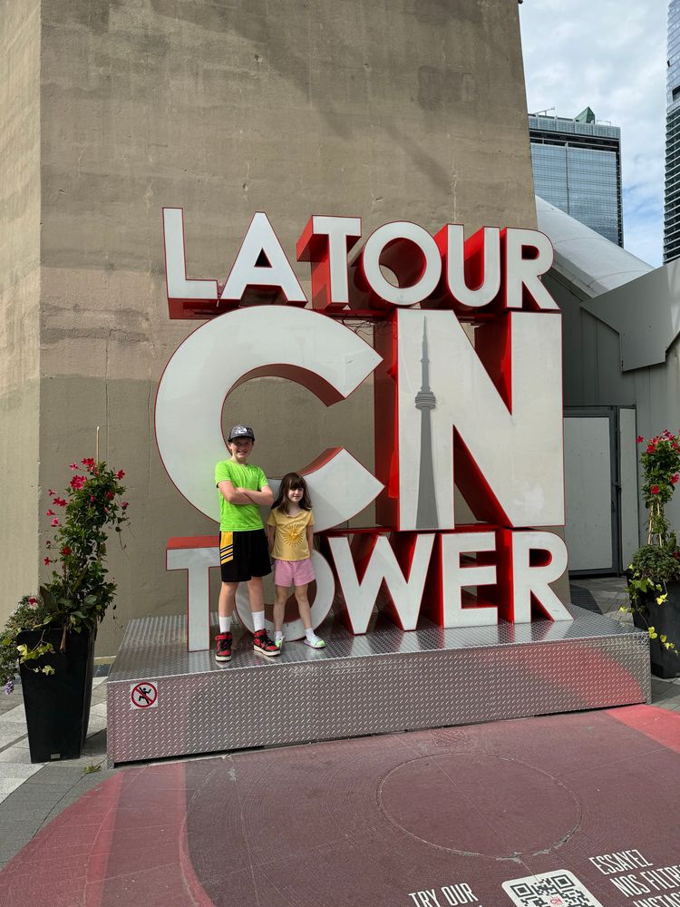 Two kids in the summer standing in front of the CN Tower sign that says La Tour CN Tower. 