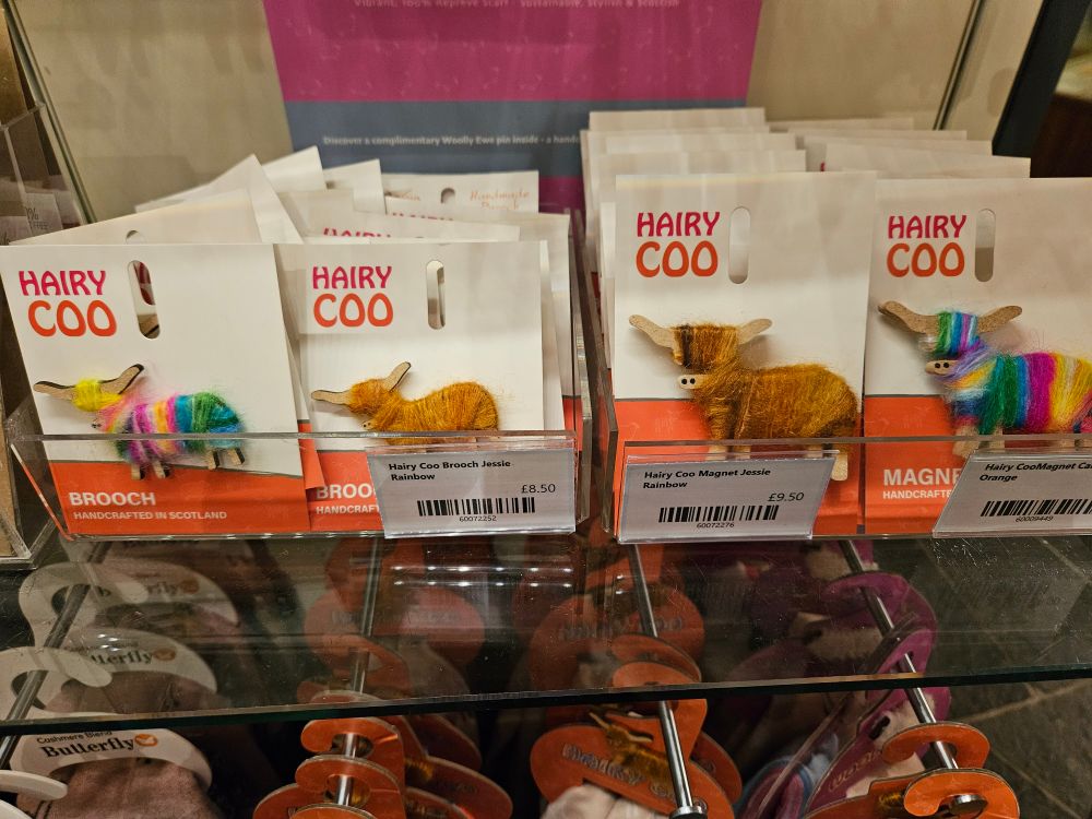 Envelopes pequenos em exibição numa prateleira de loja. Cada um deles contém um ímã de geladeira em formato de boi peludo, dois castanhos e dois com as cores do arco íris. Cada um deles traz o texto "HAIRY COO" em laranja