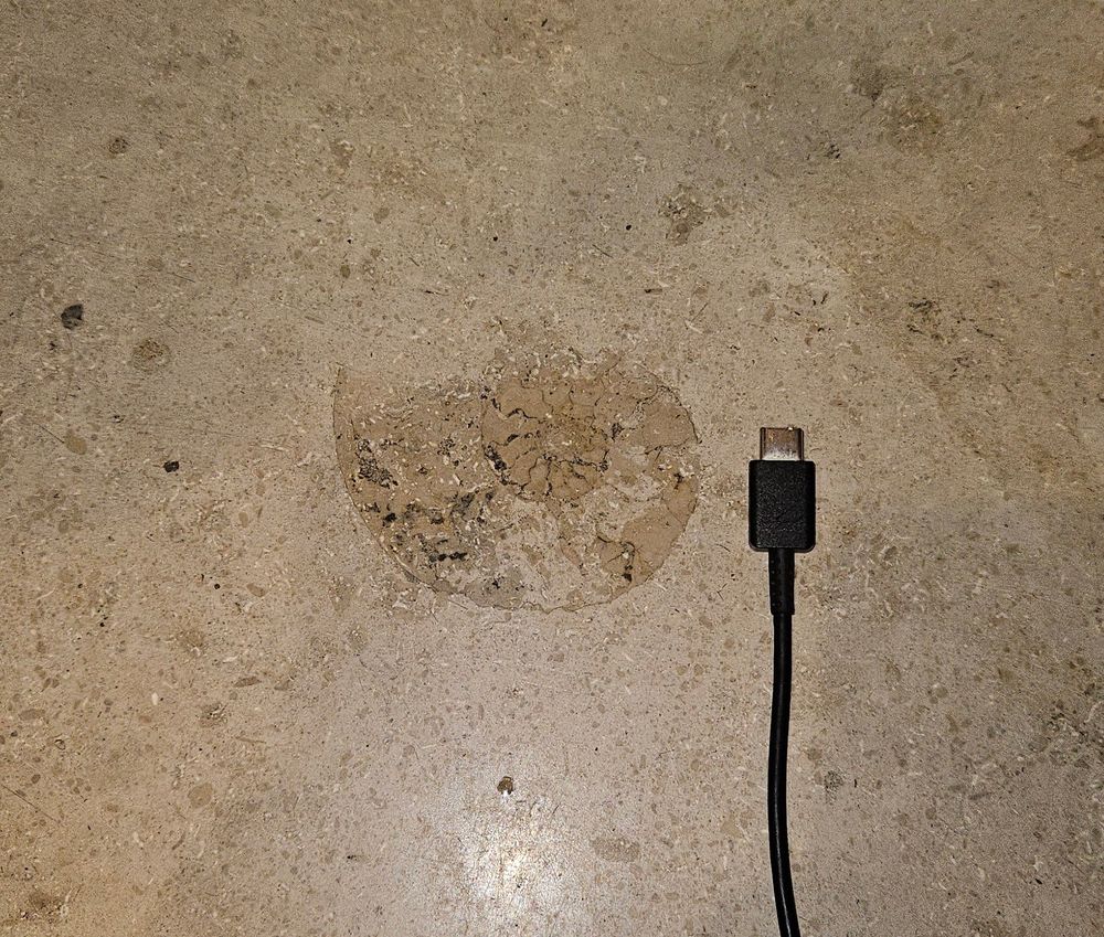 Foto do revestimento do piso, cor bege. No centro, uma impressão marrom em forma de espiral, correspondendo a um fossil de amonita. Ao lado, cabo USBc pra referência, mostrando que a imagem tem uns 10cm