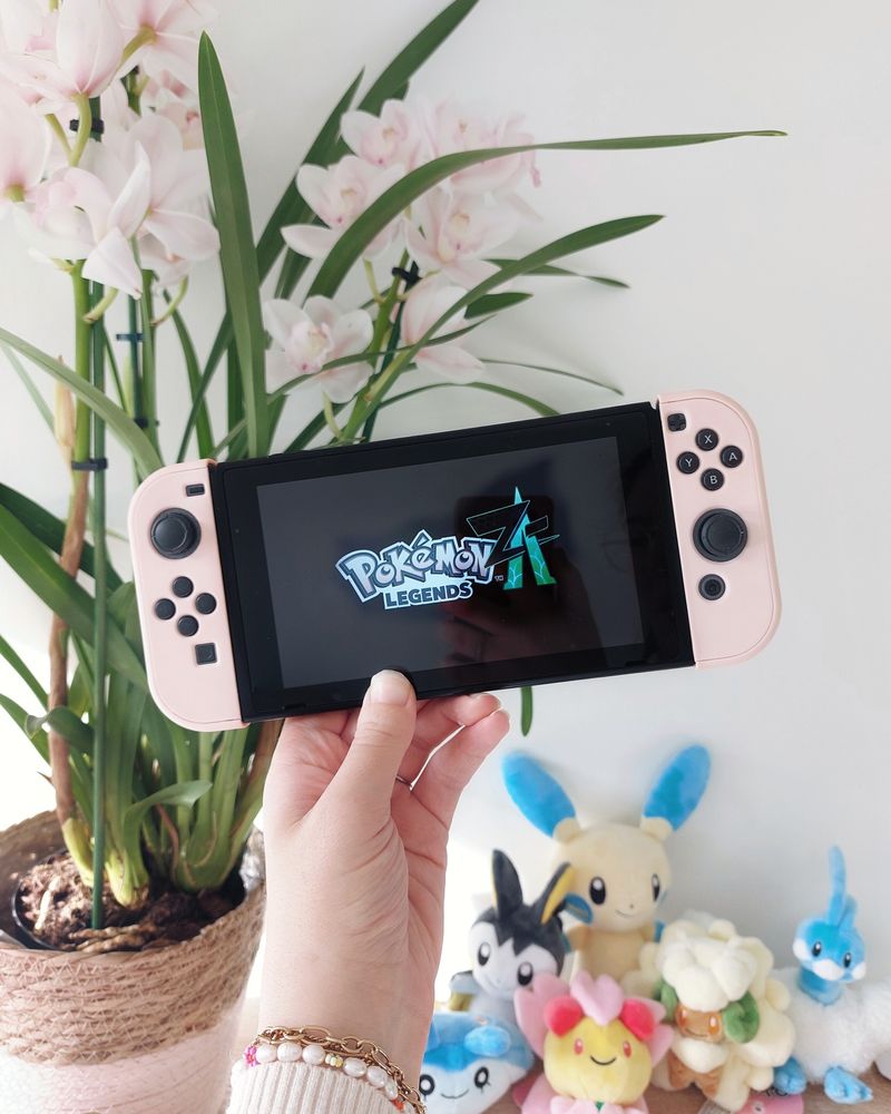 A picture of my hand holding up my Nintendo Switch in front of a white wall, with an orchid plant on the left and several Pokémon plushies on the right. On the Nintendo Switch screen, the logo of Pokémon Legends ZA is displayed. The Nintendo Switch joycons have pastel pink cases around them.