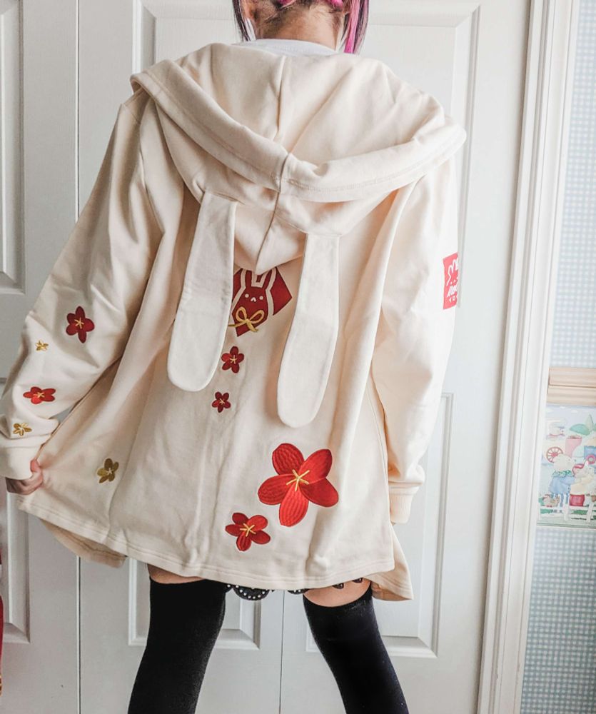 photo of a person wearing a long off-white cardigan embroidered with red flowers and bunny ears on the hood, from behind