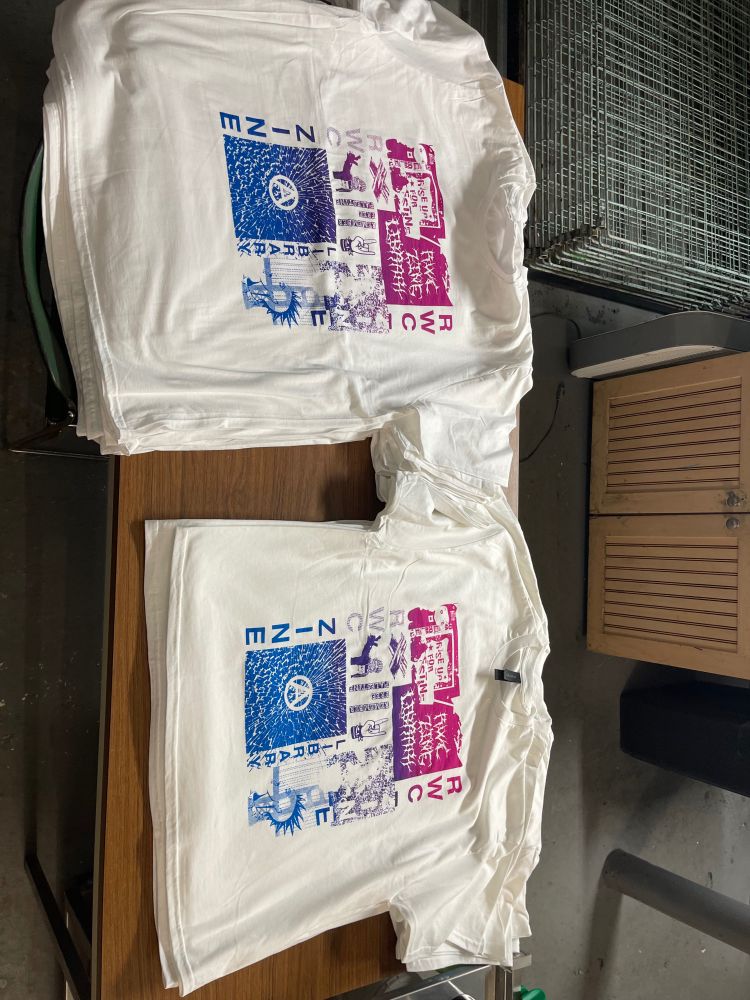 There are two piles of white shirts in a screenprinting studio. The shirts are on a brown table with a printmaking drying rack and cabinet in the background. The shirts have the Redwood City Zine Library collage design printed on them in a gradient from hot pink to cyan. The colors blend to purple in the middle. The design includes phrases like RWC ZINE LIBRARY and RWC ZINE, the anarchist symbol, free palestine, and a cartoon of a punk. The shirts are piled and ready to be shipped!