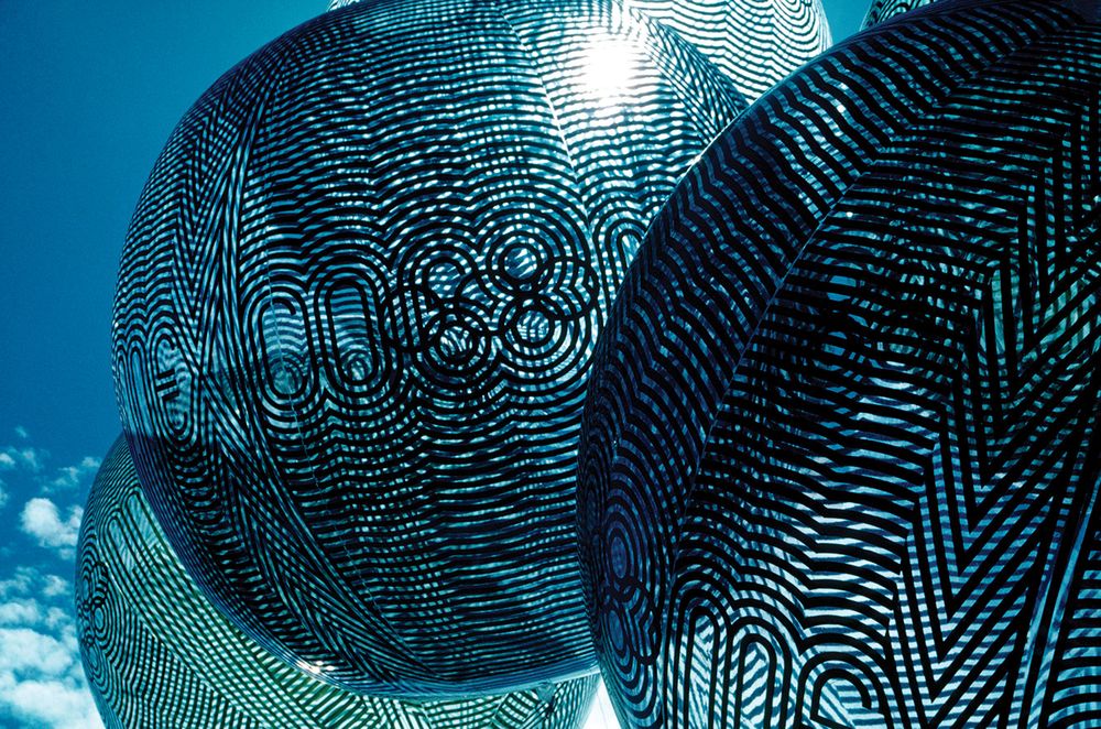 Clear balloons printed with a black graphic pattern derived from the Mexico 68 logo float in front of a blue sky.